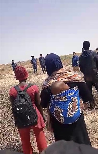 Un groupe de migrantes et de demandeureuses d’asile noires de plusieurs nationalits africaines, dont une femme et son bb, bloques dans le dsert depuis des jours aprs avoir t expulses de Tunisie, debout dans la zone tampon  la frontire entre la Tunisie et la Libye, face  une quipe de journalistes d’Al Jazeera et  des soldats libyens, le 11 juillet 2023.   2023 Priv