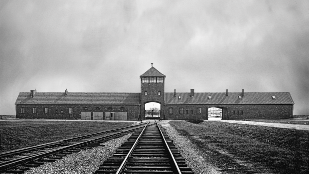 Image d'archives en noir et blanc montrant au premier plan des voies ferres menant au loin vers l'entre du camp d'Auschwitz.